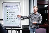 Ein Mann präsentiert vor einem Flipchart in einem Konferenzraum. Das Flipchart zeigt handschriftliche Notizen und Diagramme. Die Teilnehmer beobachten aufmerksam die Präsentation.